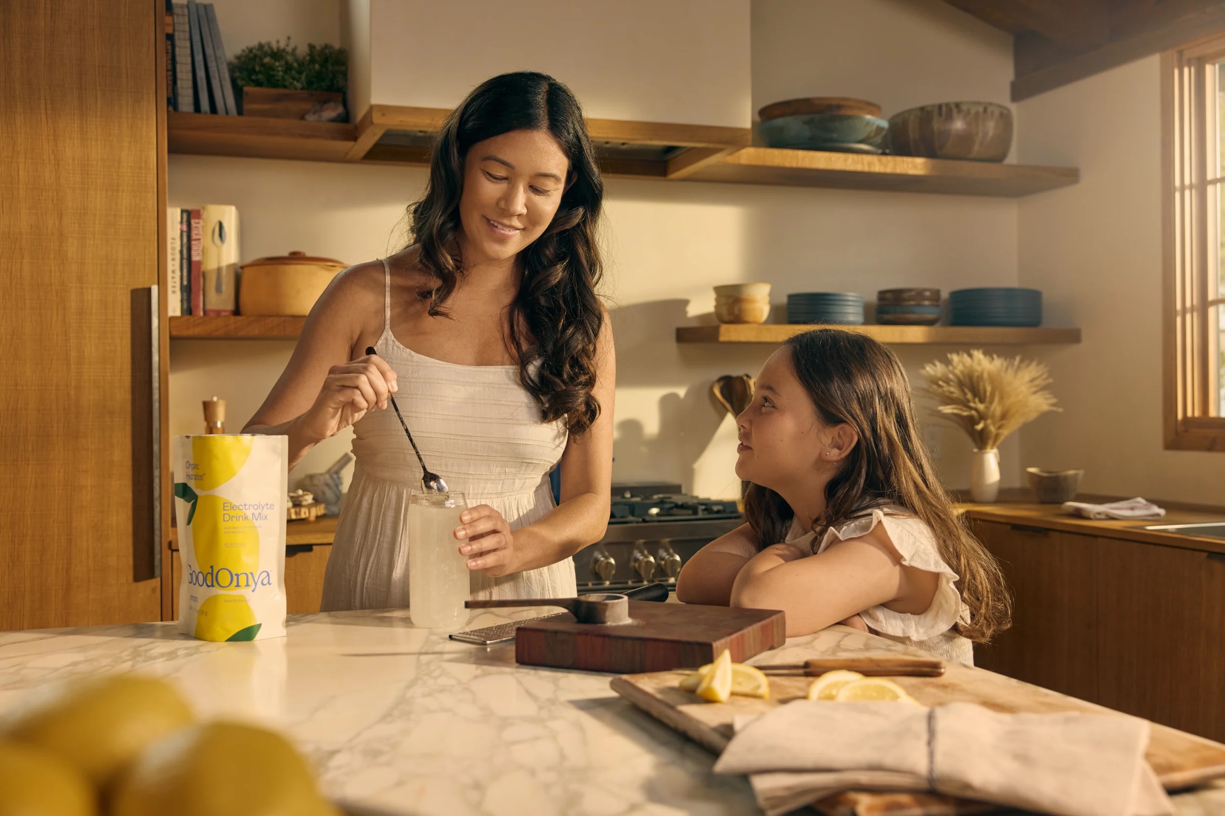 A woman stirs a drink in a glass jar while a young girl watches her at a kitchen counter with lemons, a napkin, and a lemonade drink mix.
