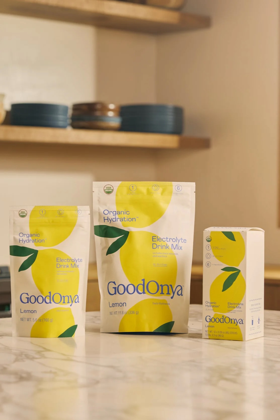 Three GoodOnya Lemon Electrolyte Drink Mix packages are displayed on a kitchen counter, including two pouches and one box, with shelves and dishes in the background.