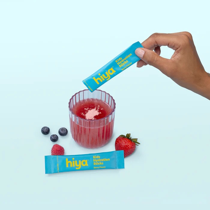 A hand pours a Hiya Kids Hydration Stick into a glass of red liquid, with a strawberry and blueberries placed nearby on a light blue background.