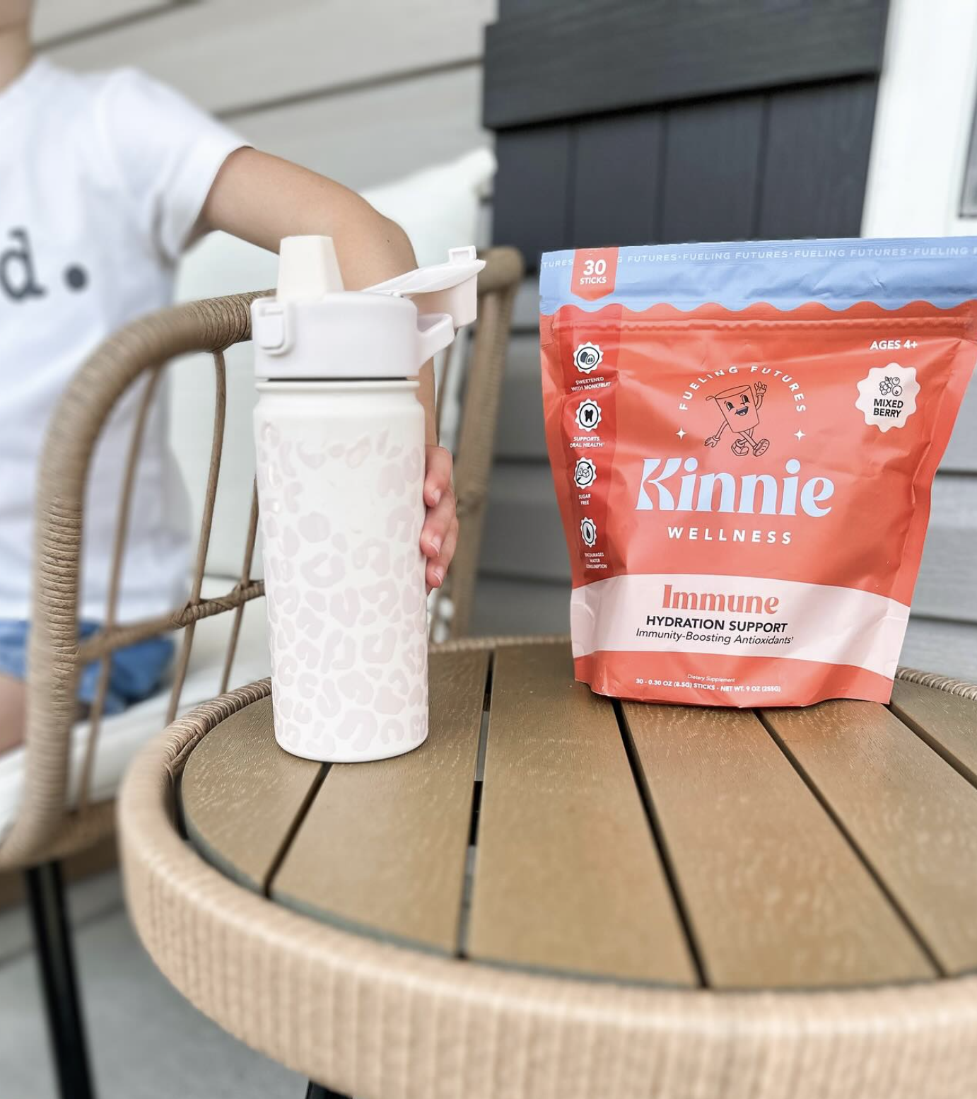 A person sits on a chair next to a table with a white water bottle and a Kinnie Wellness Immune Hydration Support drink mix bag.