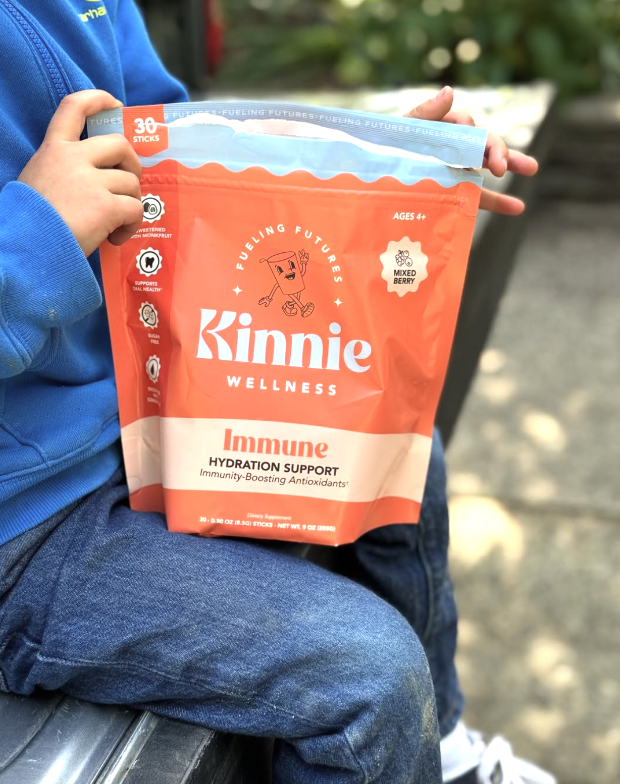 A child holding a large orange and white bag of Kinnie Wellness Immune hydration support powder sticks while sitting outdoors.