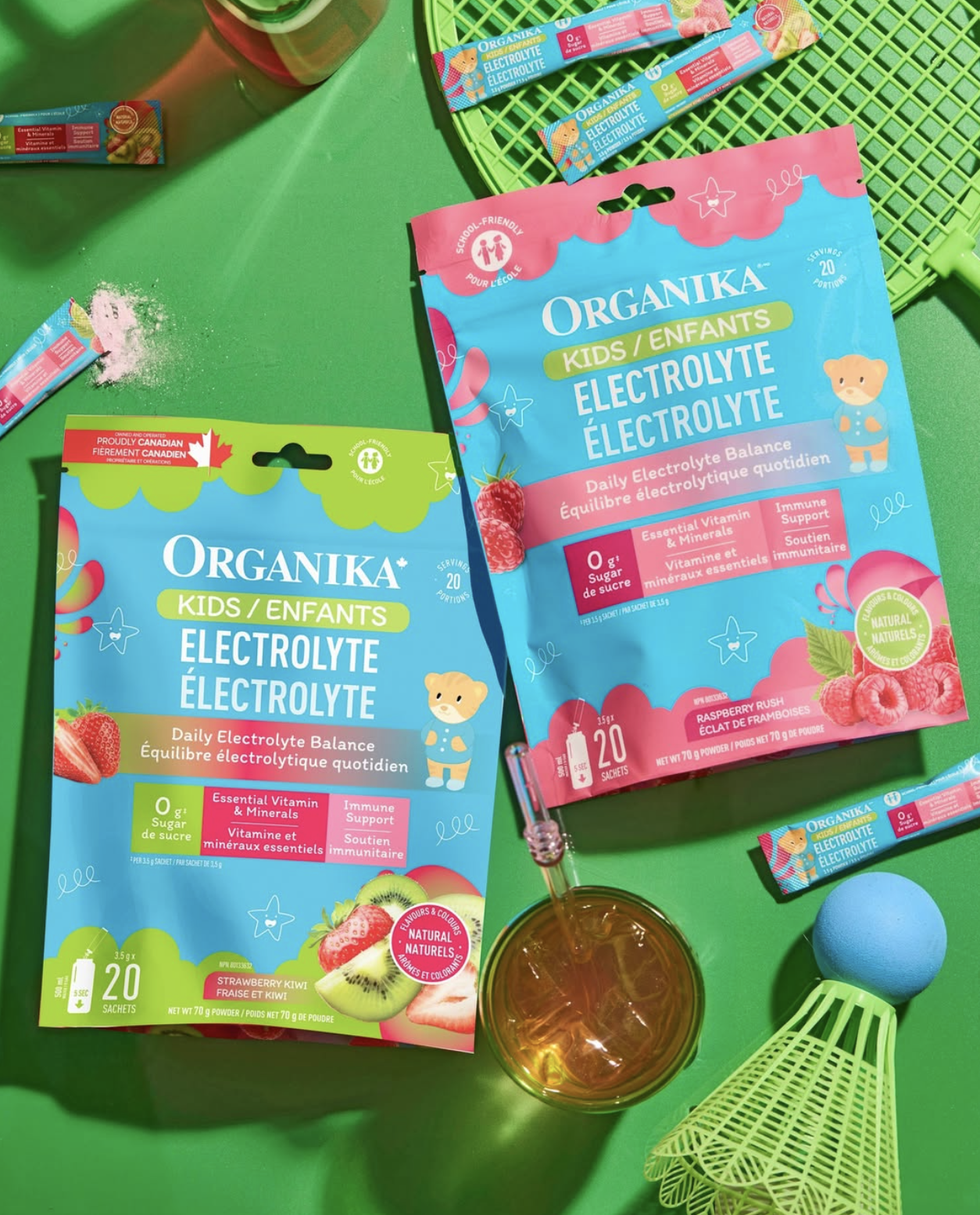 Two packets of Organika Kids Electrolyte powder, raspberry and tropical punch flavors, displayed on a green surface with a badminton racket, birdie, drink, and powder sticks.