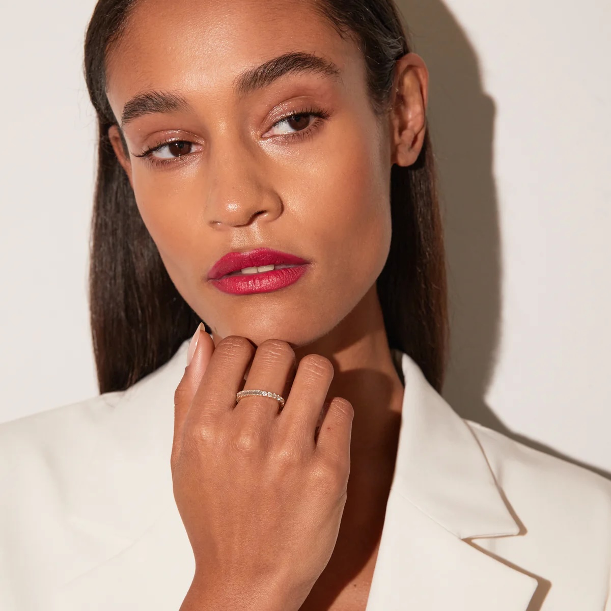 A woman in a white blazer with straight hair looks to the side, resting her hand with a ring on her chin.