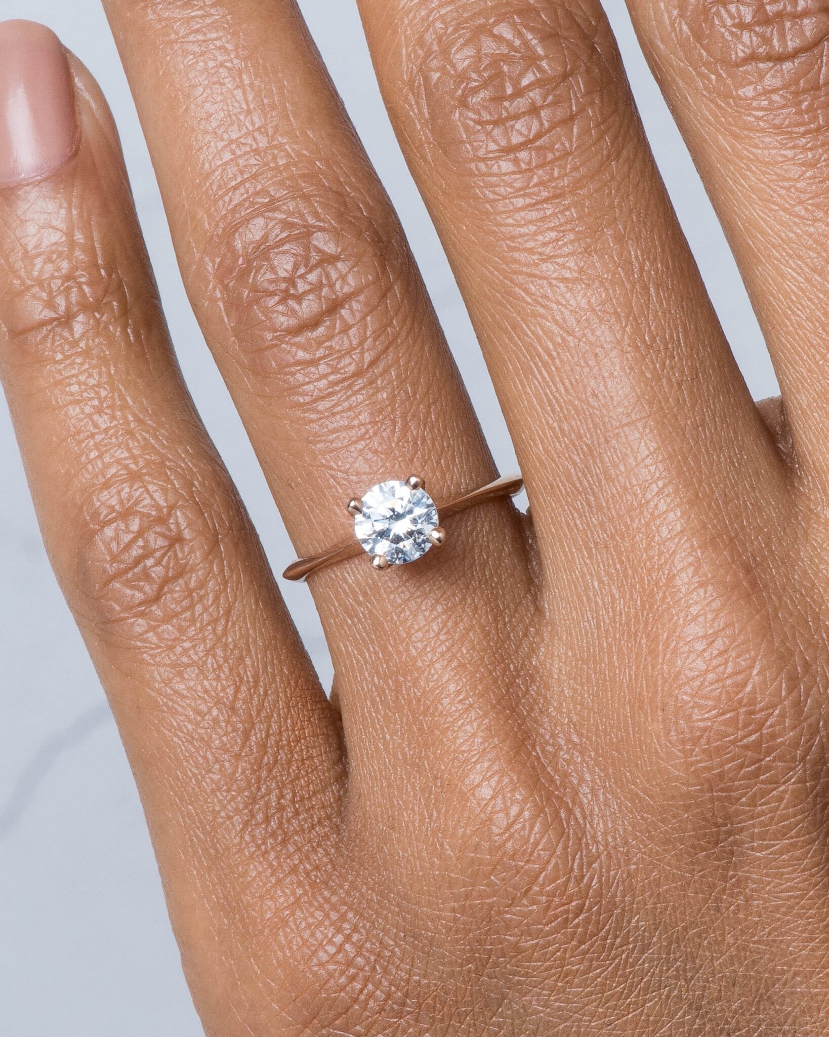 A hand with a solitaire diamond ring on the ring finger, featuring a thin band and a round, brilliant-cut diamond set in a simple prong setting.