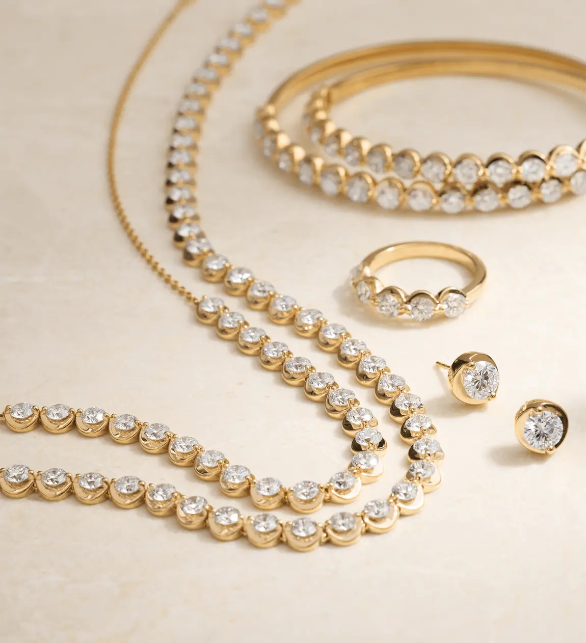 A set of gold jewelry featuring two necklaces, a bracelet, a ring, and a pair of stud earrings, all adorned with round clear gemstones, displayed on a beige surface.