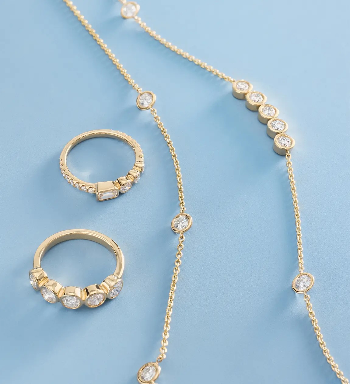 Two gold rings and a gold necklace with clear gemstones are arranged on a light blue surface.