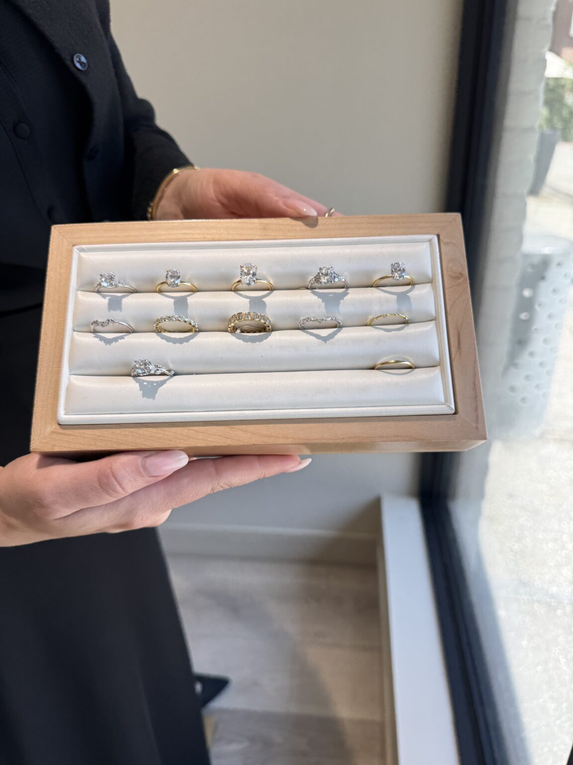 Person holding a display box with twelve assorted rings, including diamond and gold designs, near a window in a jewelry store.