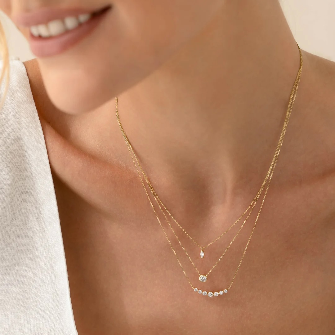 A woman wearing a white top and three layered gold necklaces with small pendants, including pearls and gemstones.
