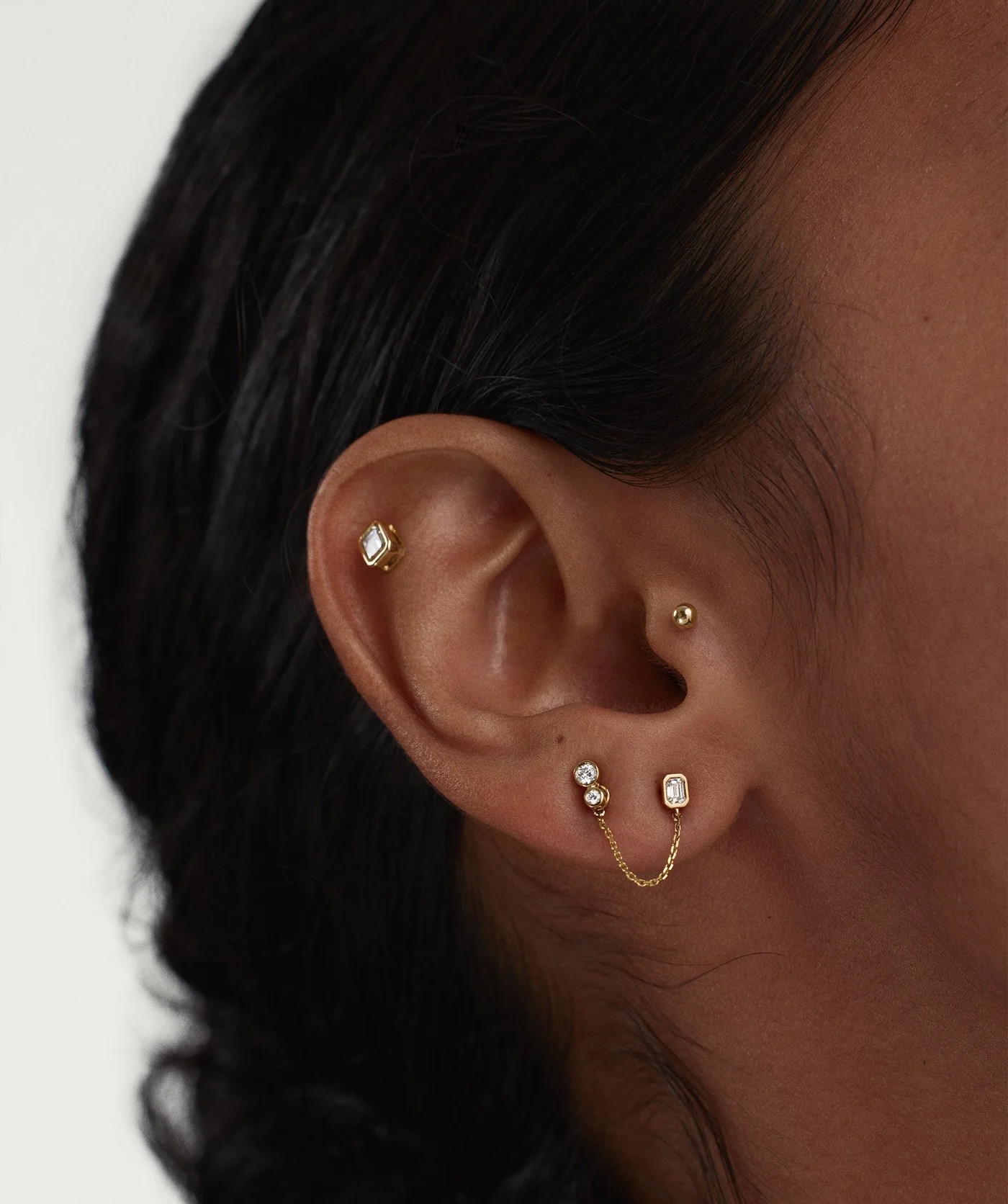 Close-up of an ear with four piercings, featuring three gold studs and one double stud connected by a delicate gold chain.