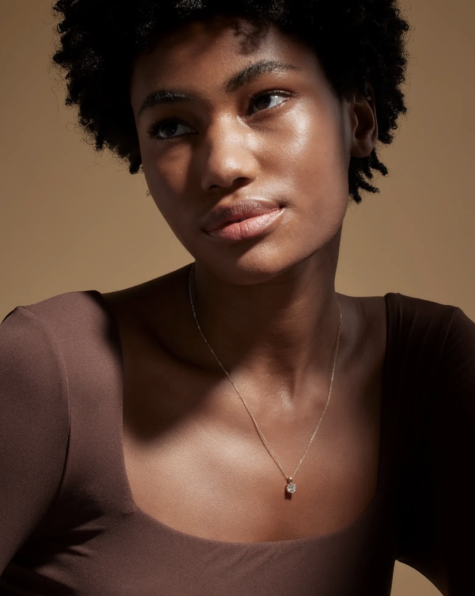 A woman wearing a brown top and a delicate necklace with a pendant poses against a plain, warm-toned background.
