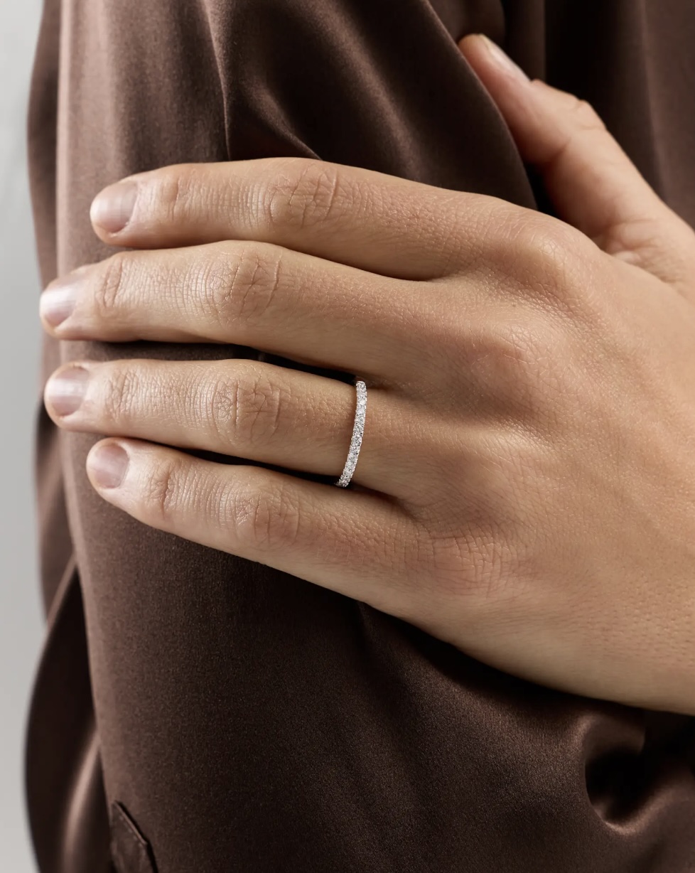 A hand with neatly trimmed nails rests on a brown satin fabric, wearing a thin, diamond-studded band on the ring finger.
