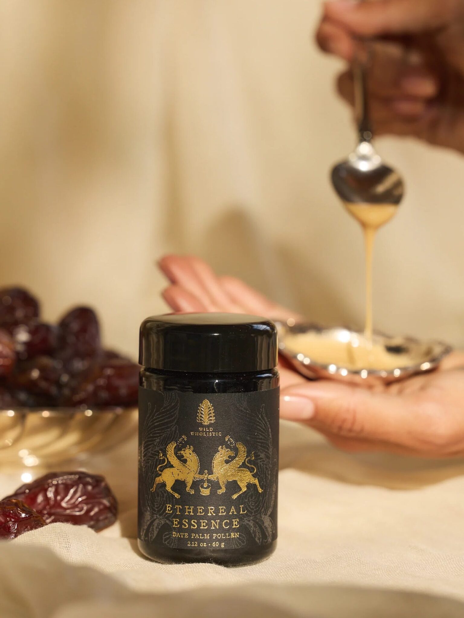 A jar labeled "Ethereal Essence Date Palm Polen" sits on a surface near dates, while a hand drizzles a spoonful of a creamy substance into a small dish.