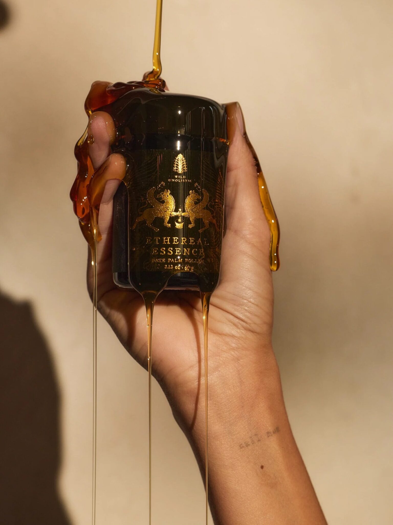 A hand holds a dark amber jar labeled "Ethereal Essence" as golden honey or syrup drips over the fingers and jar.