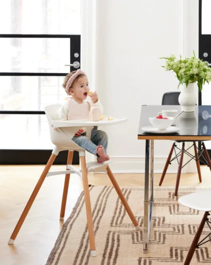 A toddler sits in a high chair eating at a table in a bright dining room with a vase of flowers and bowls on the table.