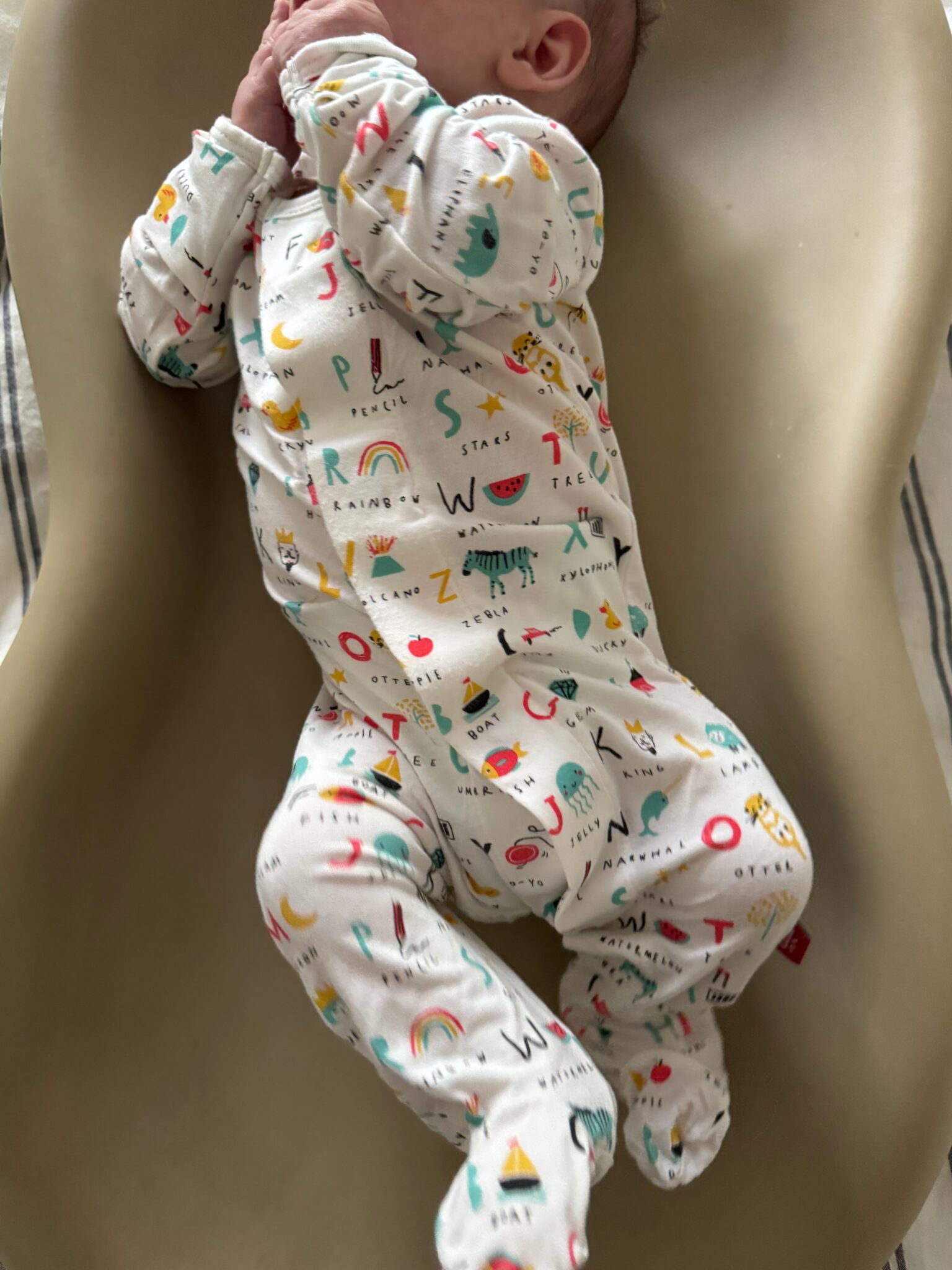 Baby in a white alphabet-themed onesie lies on a beige surface with hands covering their face.
