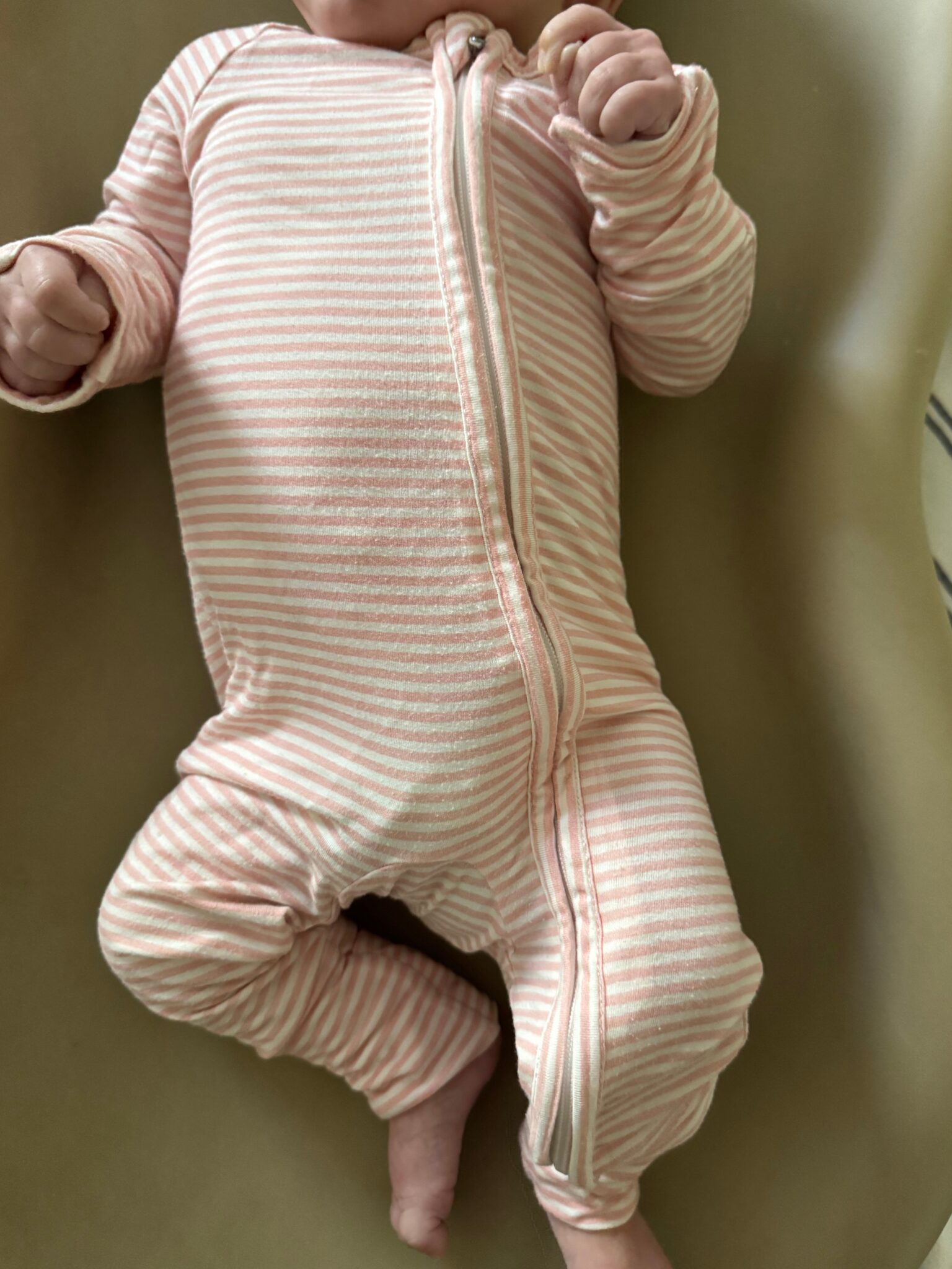 Baby wearing a light pink and white striped zip-up onesie, lying on a beige surface with only the lower face and body visible.