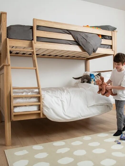 A young boy stands beside a wooden bunk bed holding two stuffed animals, with toys and a book on a polka dot rug in a bright, tidy bedroom.