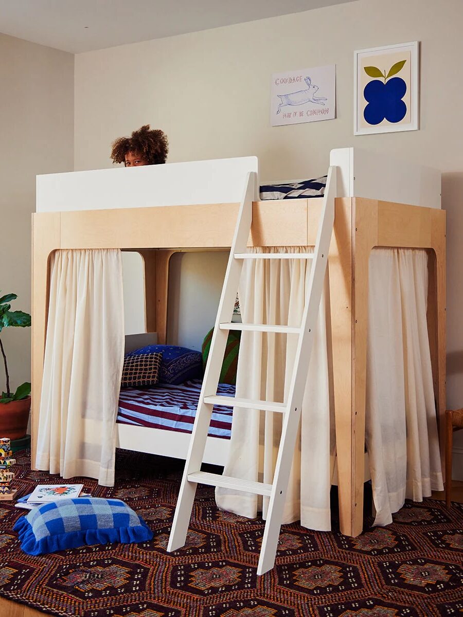 A modern wooden bunk bed with a white ladder and curtains on the bottom bunk stands on a patterned rug; a child is partially visible on the top bunk.