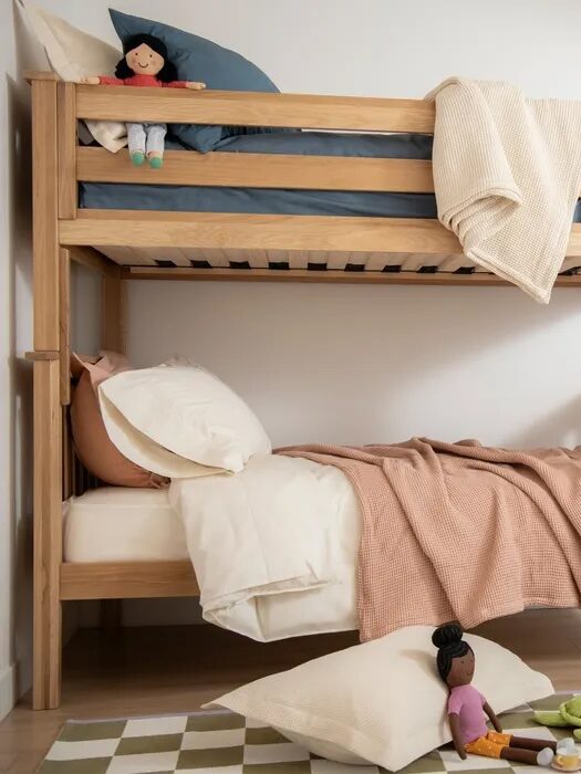 Wooden bunk bed with blue and beige bedding, dolls on both beds and floor, and a beige and green checkered rug in a children's room.