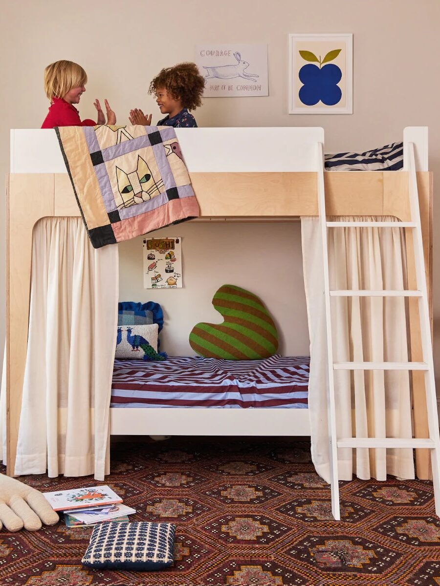 Two children play on the top bunk of a wooden bunk bed with a ladder, patterned bedding, and curtains, in a cozy, decorated room.