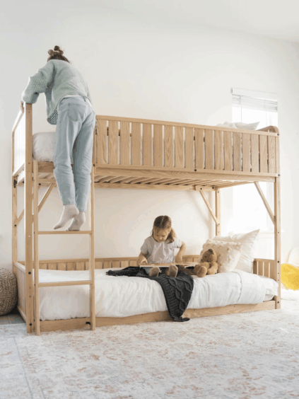 Two children use a wooden bunk bed; one climbs the ladder to the top bunk while the other sits on the lower bunk, reading a book next to a teddy bear.