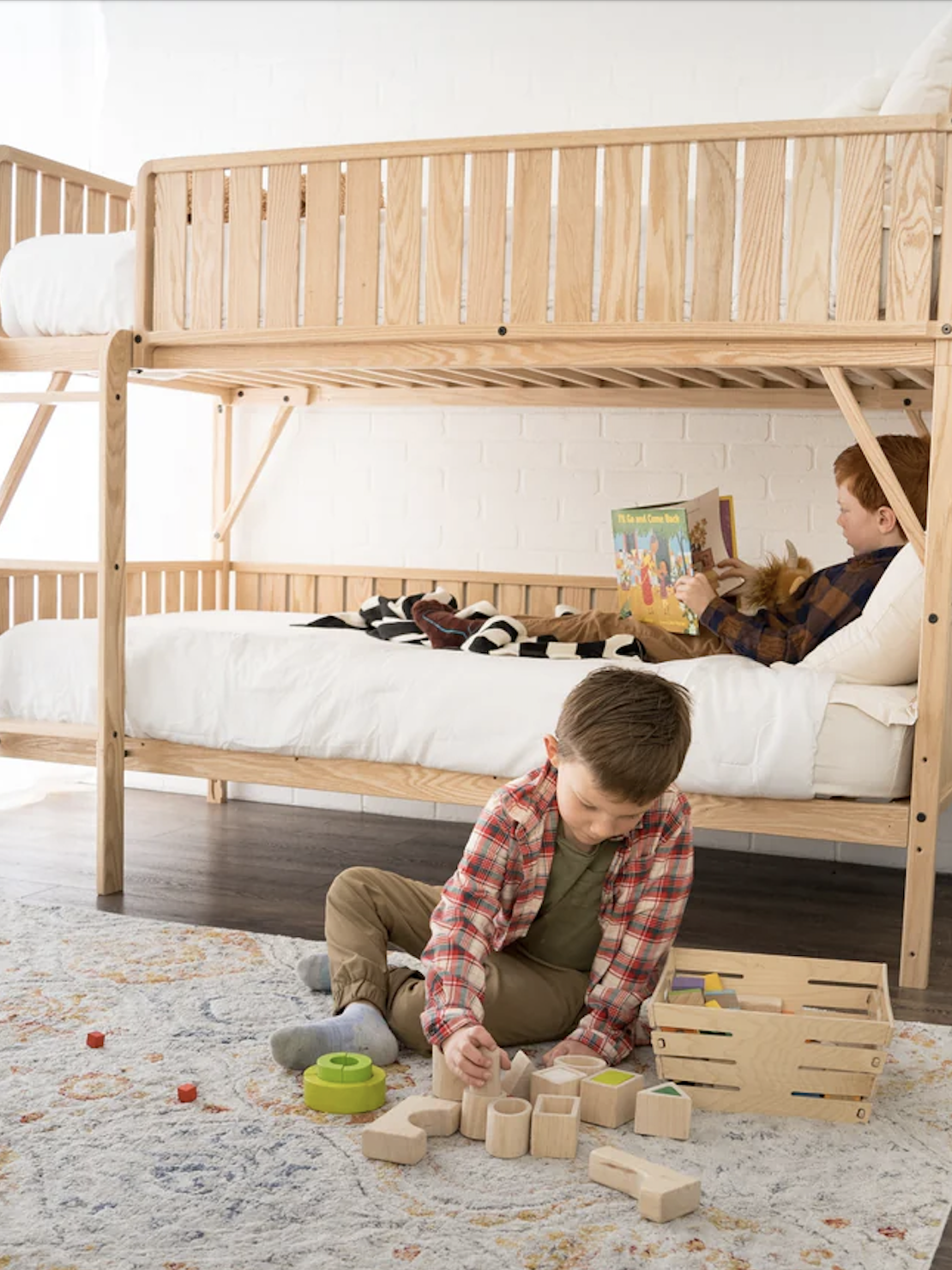 Two children are in a bedroom with wooden bunk beds; one reads a book on the lower bunk, while the other plays with wooden toys on the rug.
