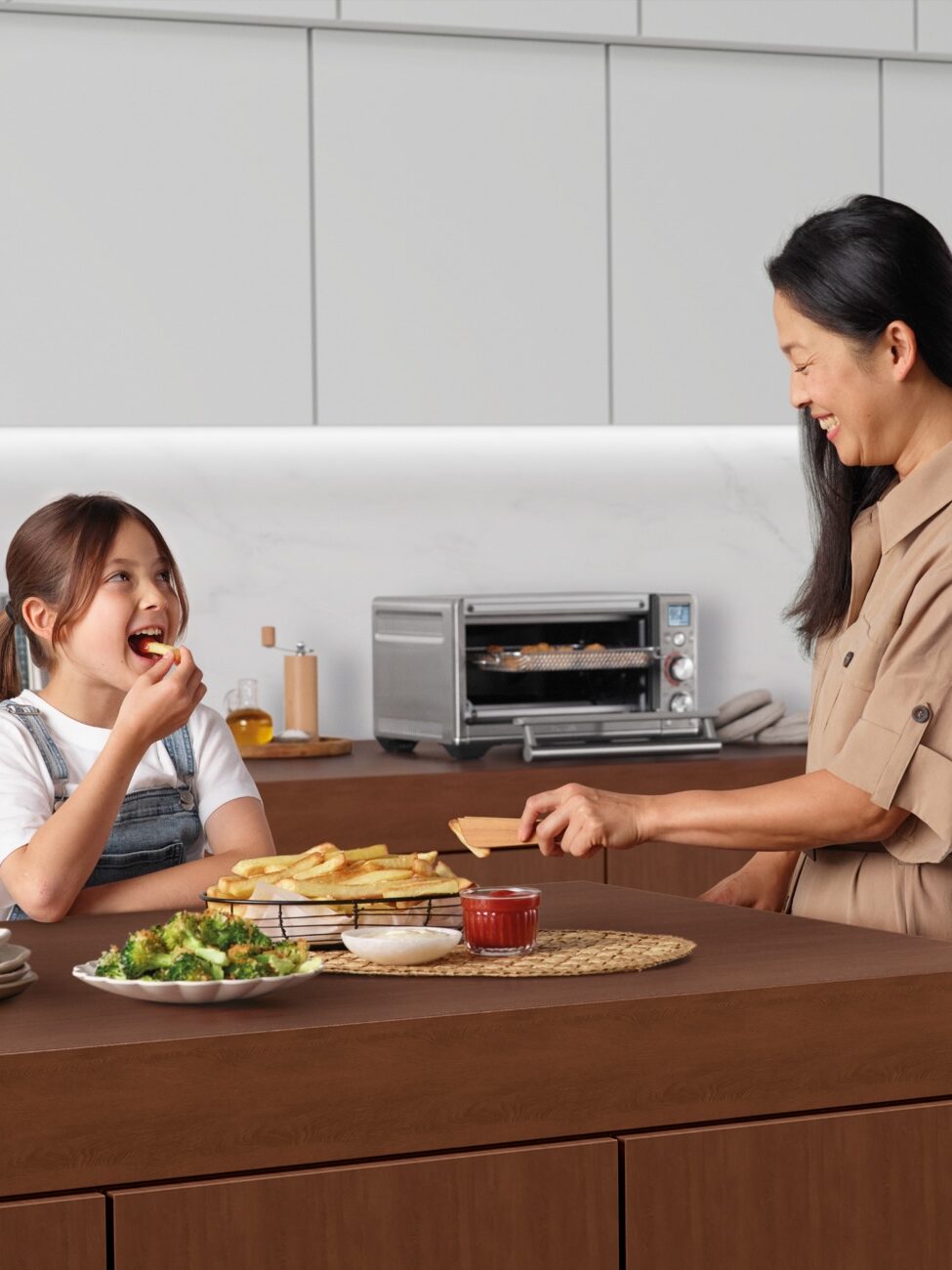 A woman serves food to a young girl in a modern kitchen. The girl sits at the counter eating, while the woman smiles and holds a plate. Food and kitchen appliances are visible.