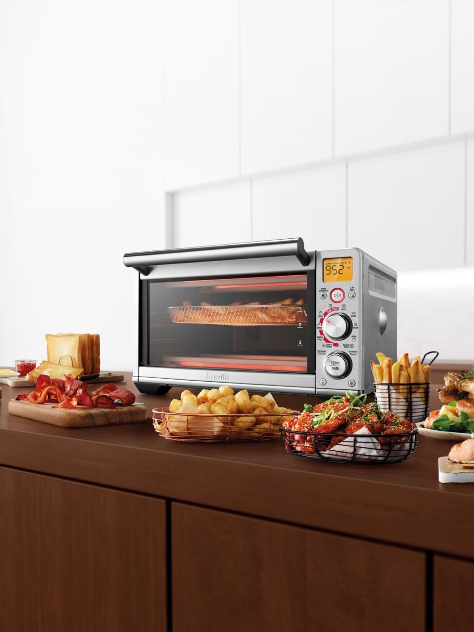 A countertop oven baking salmon with various cooked dishes including fries, chicken, and pizza arranged on a kitchen island.