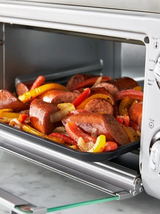 A baking tray filled with sliced sausage, red and yellow bell peppers, and onions is placed inside a countertop toaster oven.