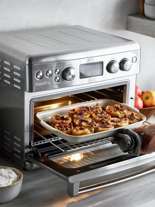 A tray of cinnamon rolls is being removed from a stainless steel countertop oven in a kitchen setting.