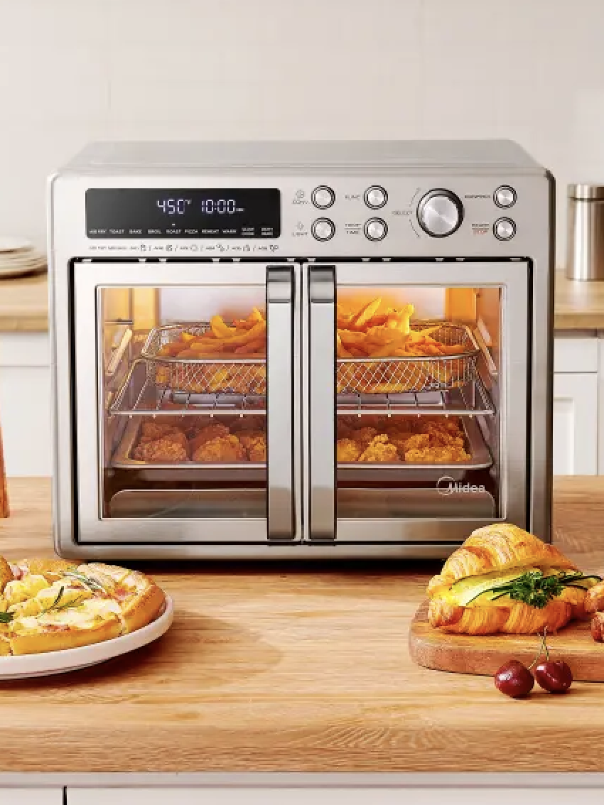 A countertop air fryer oven with double doors is cooking food inside, while croissants and a pizza slice are on plates on the wooden counter in front.