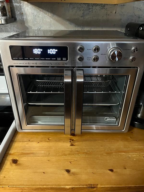 A stainless steel Midea countertop convection oven with digital display shows settings at 180°C and 1:00, sitting on a wooden kitchen counter.