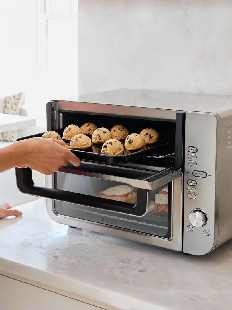 A person removes a tray of chocolate chip cookies from a countertop toaster oven in a modern kitchen.