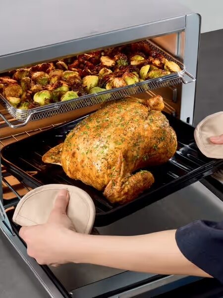 A person removes a roasted chicken from a countertop oven, with a tray of roasted Brussels sprouts visible above.