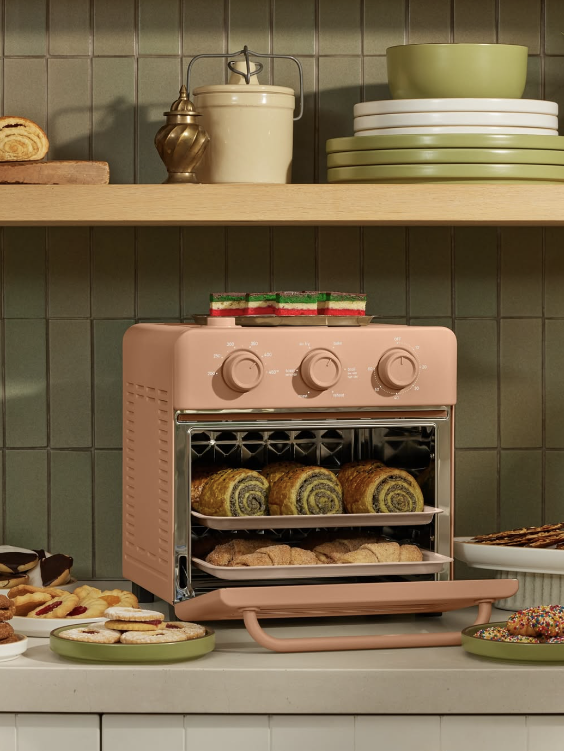 A pink countertop oven with an open door shows trays of baked goods inside, surrounded by various cookies and plates on a kitchen counter and shelves.