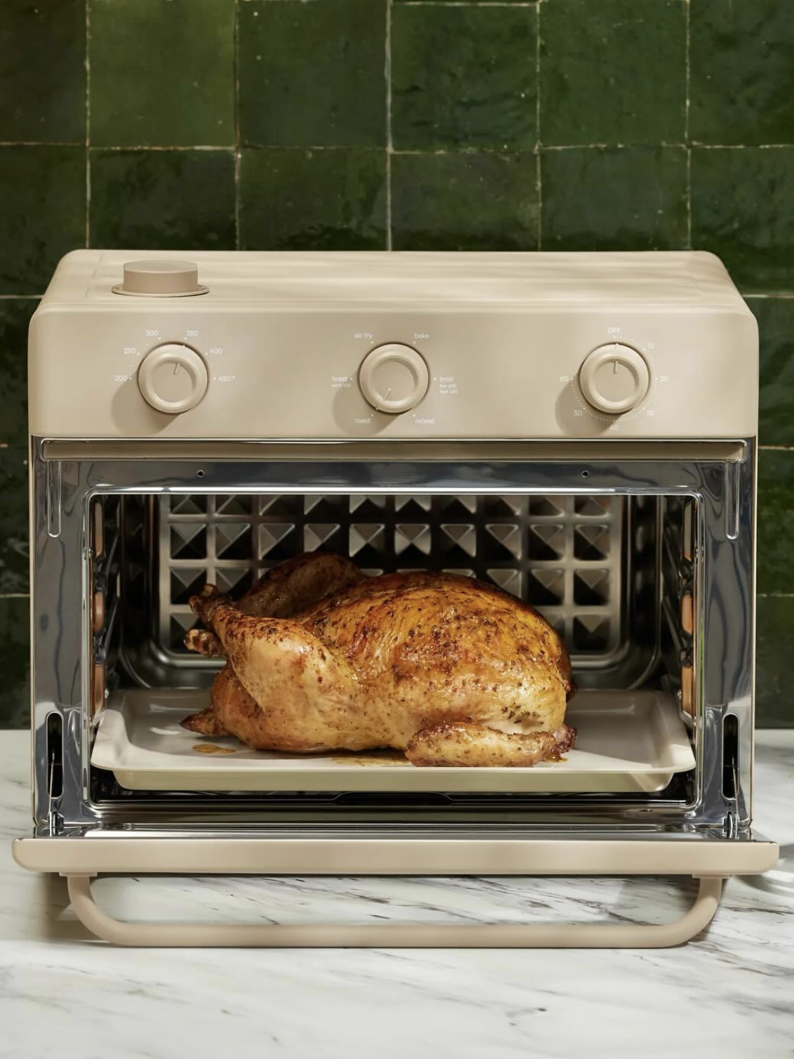 A whole roasted chicken sits on a tray inside an open beige countertop oven, with green tiled wall and marble counter in the background.