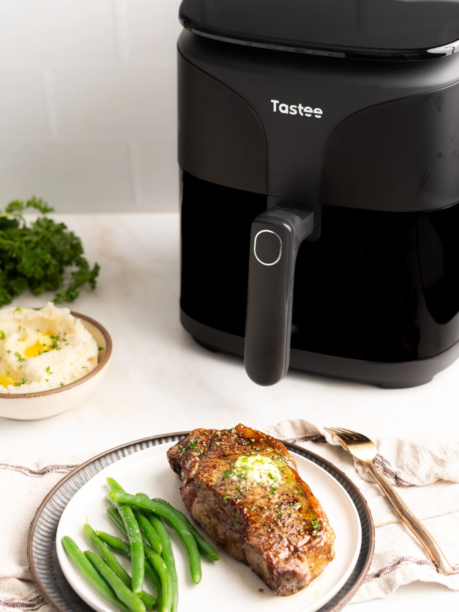 A plate with cooked steak topped with herb butter and green beans sits next to a black air fryer on a white countertop. Mashed potatoes are in a bowl in the background.