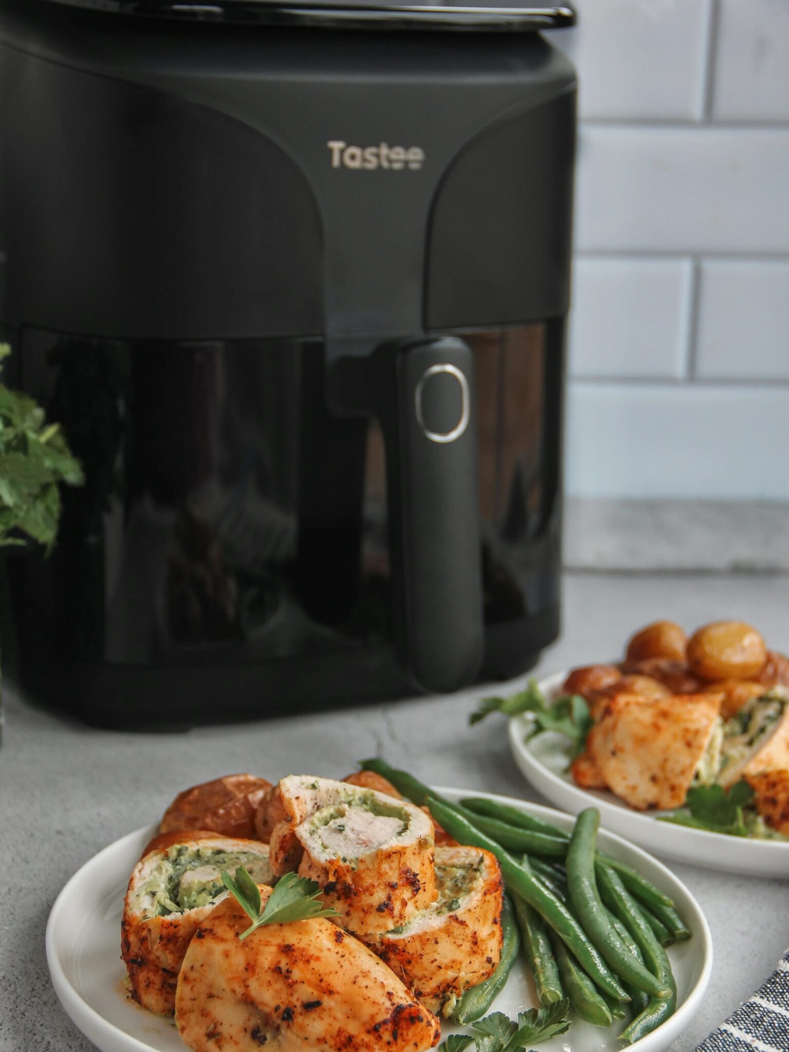 Sliced stuffed chicken breast with green beans and roasted potatoes on a white plate in front of a black air fryer.