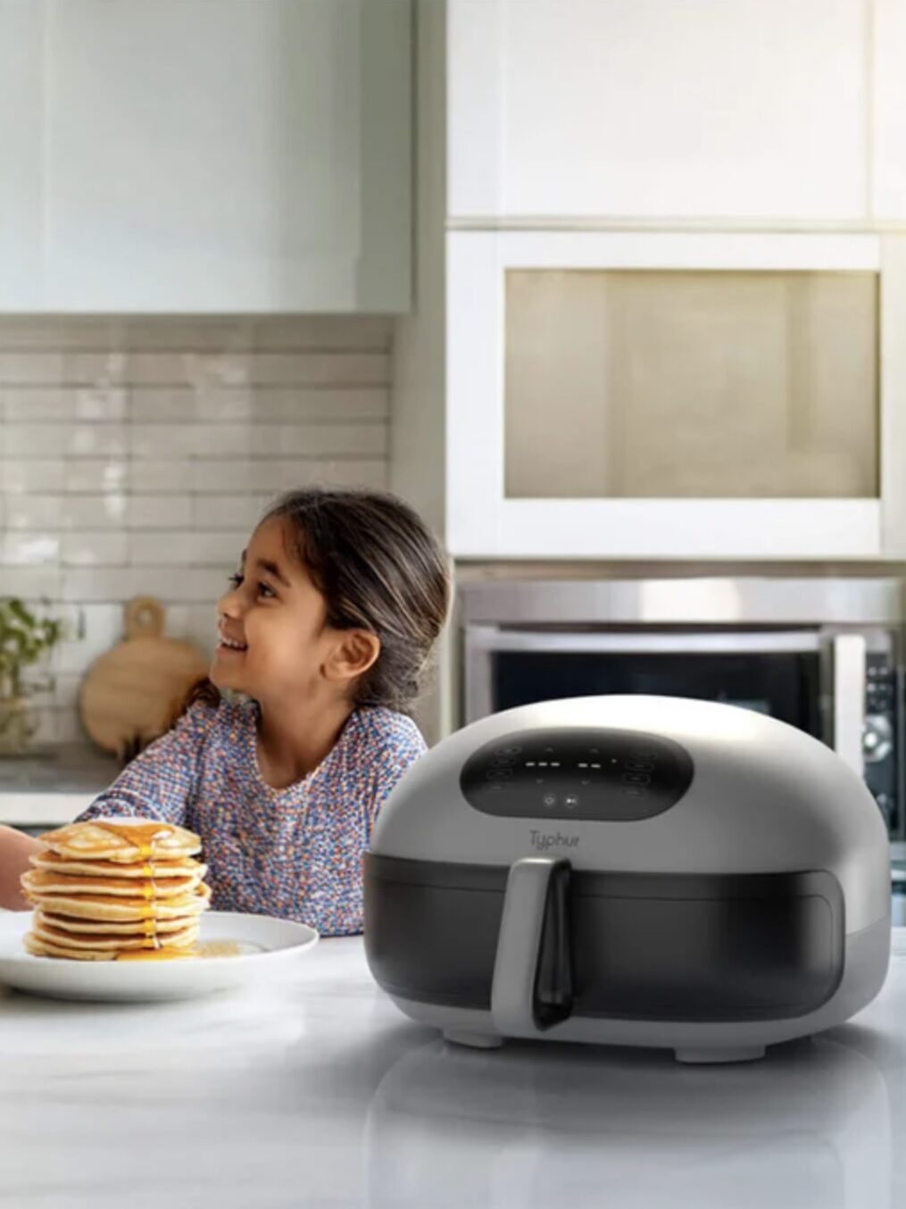 A woman and a young girl smile at each other in a kitchen while preparing a stack of pancakes; an air fryer sits on the counter in the foreground.
