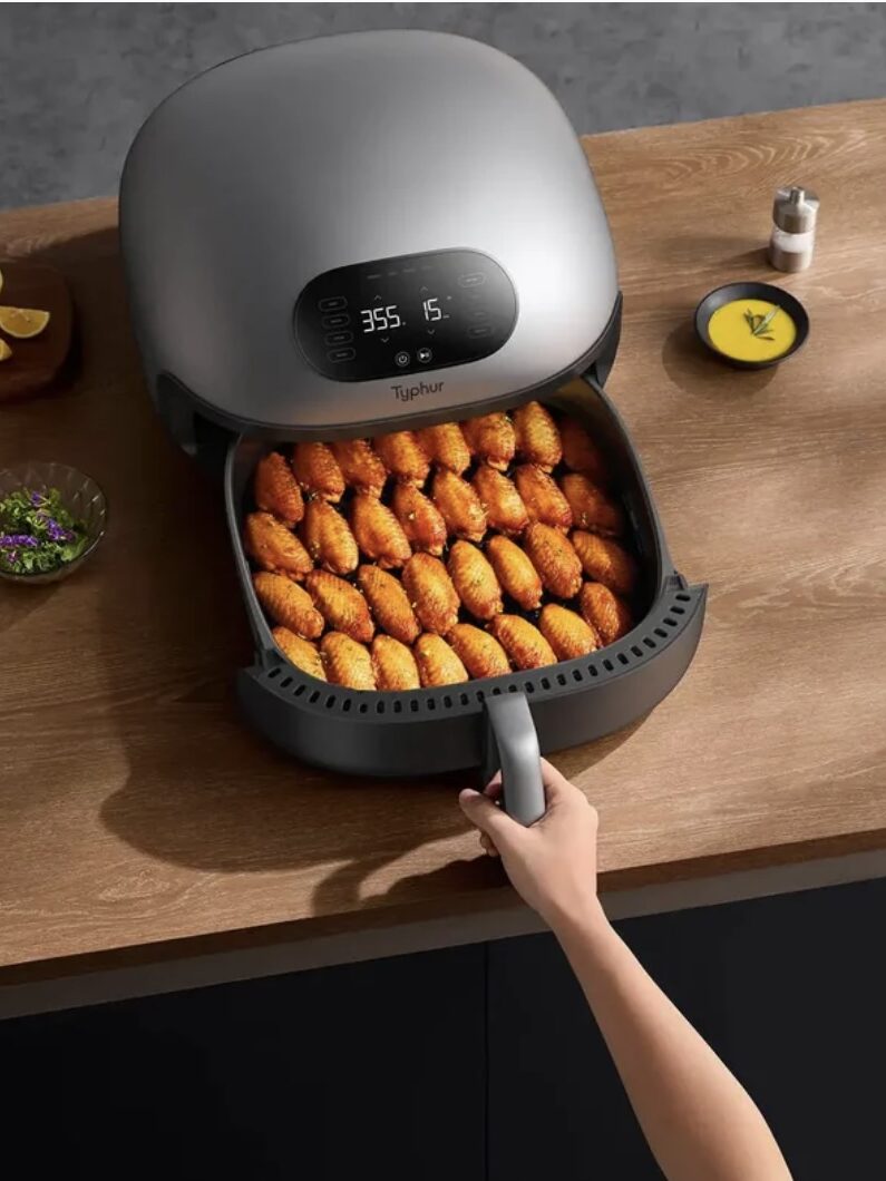 A person pulls out a tray of cooked, seasoned chicken wings from a gray air fryer on a wooden countertop.
