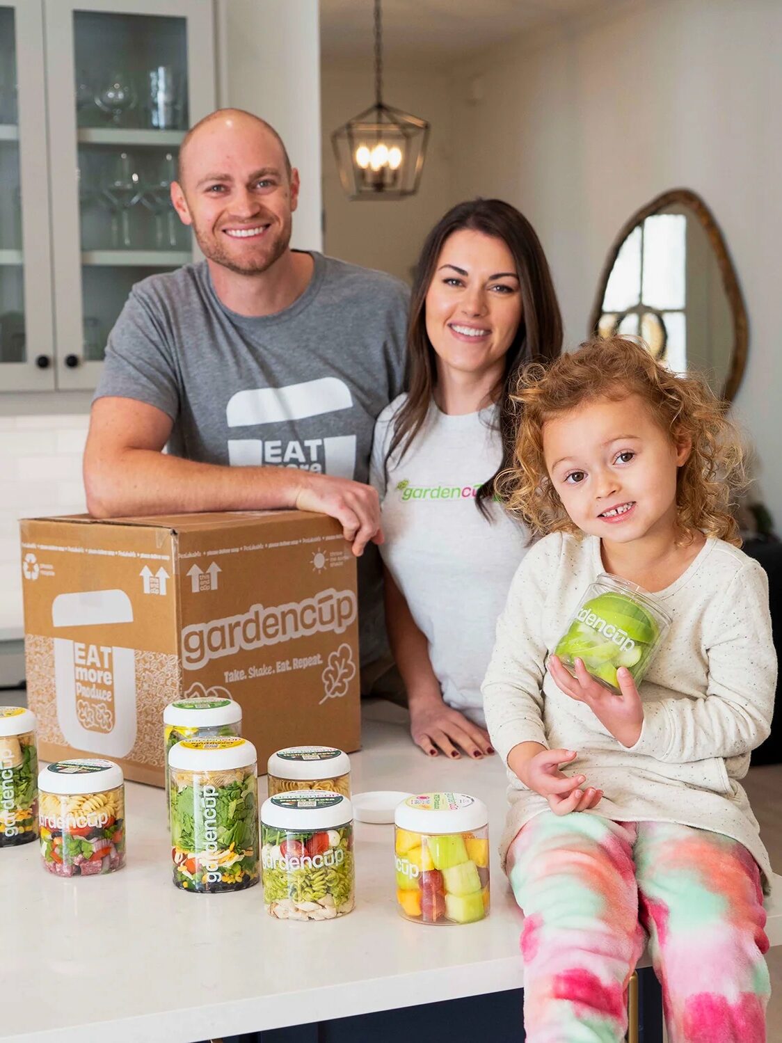 Two adults and a child pose in a kitchen with a variety of Garden Cup salad jars and a branded box arranged in front of them on the counter.
