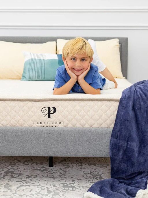 A young boy lies on his stomach on a neatly made bed with a beige and gray mattress labeled "PlushBeds" in a bright, modern bedroom.