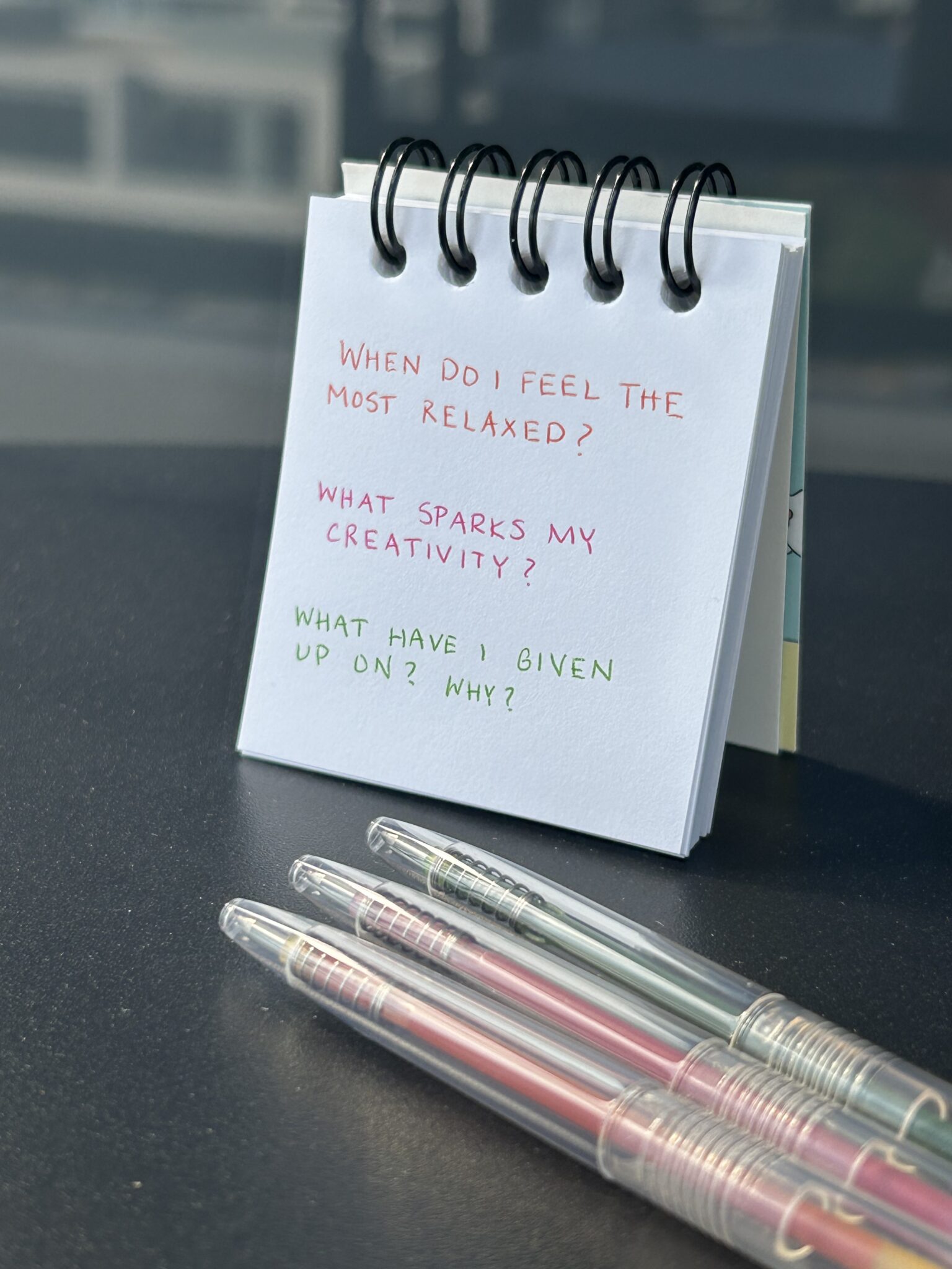 A small notepad with handwritten reflective questions in colored ink stands upright on a desk next to three pastel gel pens.