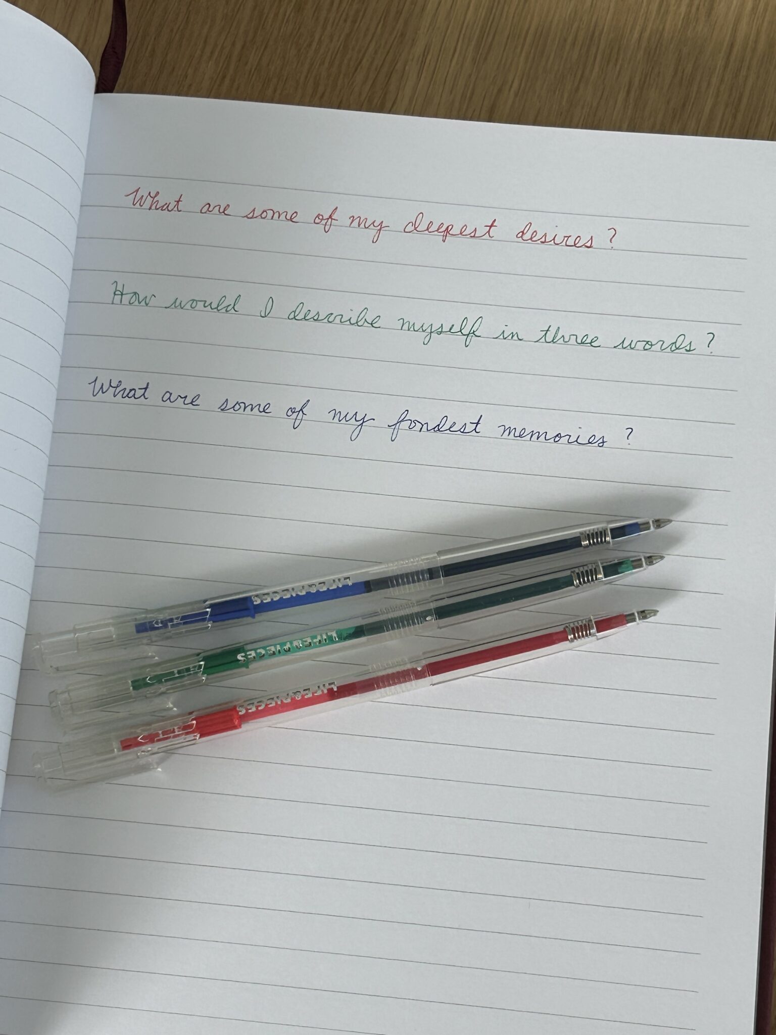 An open notebook with three handwritten prompts in red, green, and blue ink, and three matching colored pens placed across the page.