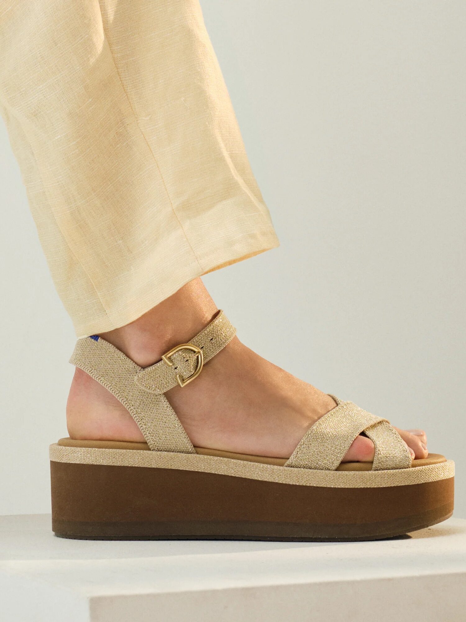 A person wearing light beige pants and beige platform sandals with a buckle stands on a white surface.