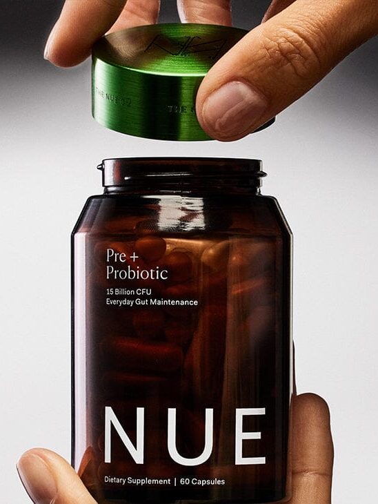 A hand opens a brown bottle labeled "NUE Pre+Probiotic" containing 60 capsules, with a green lid being lifted off.