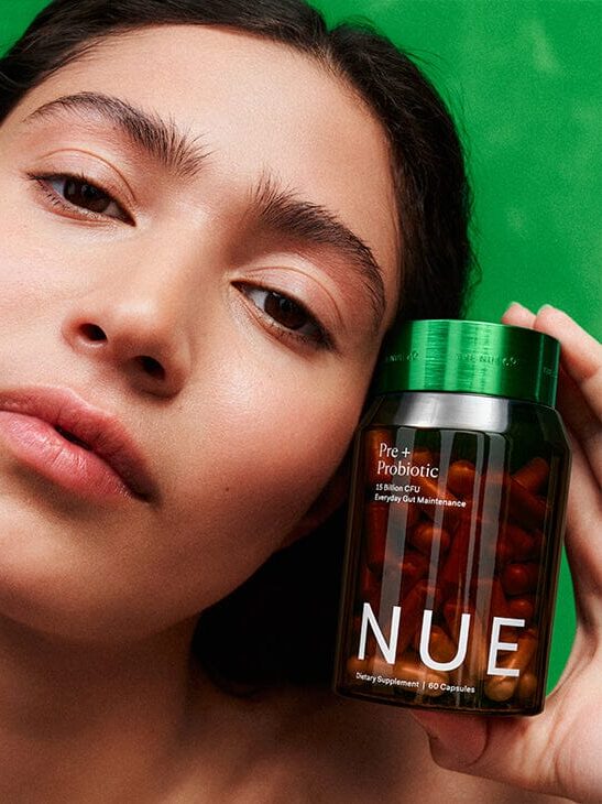 A person holds a jar labeled "NUE Pre + Probiotic" with red capsules inside, against a solid green background.