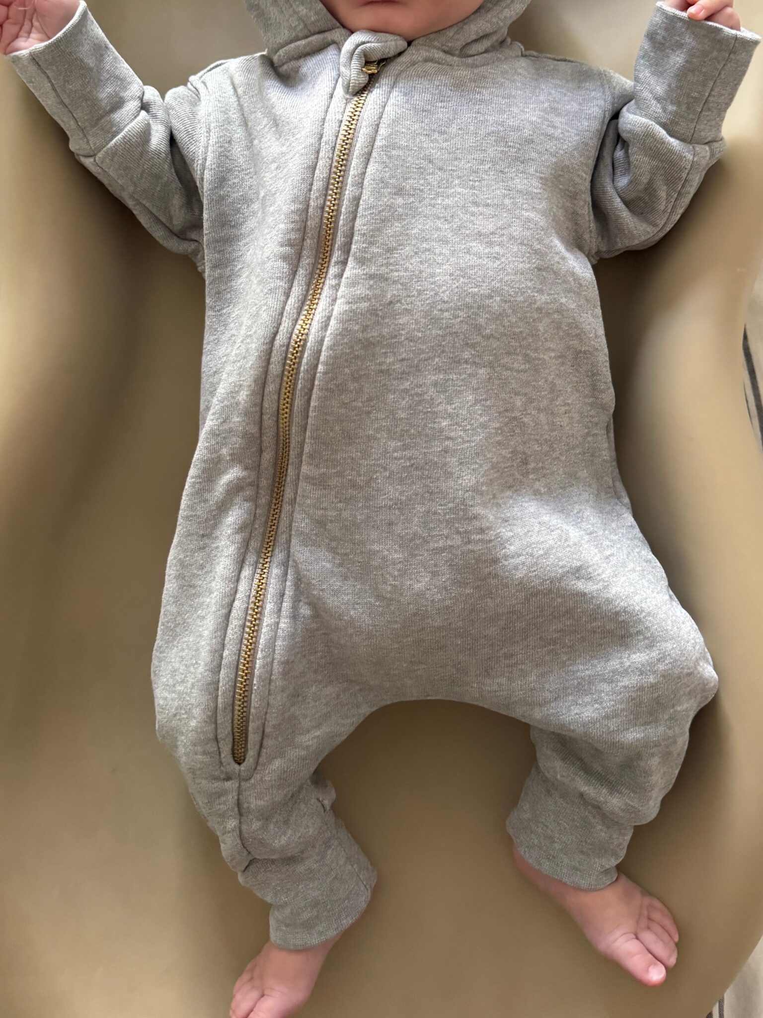 A baby wearing a light gray hooded onesie with a gold zipper, lying on a beige surface with bare feet visible.