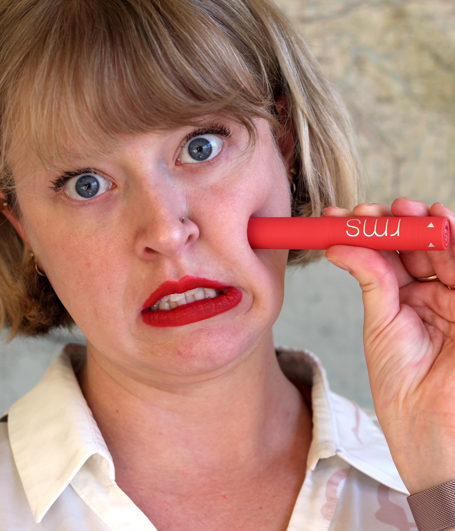 A person with light hair and red lipstick presses a red RMS Beauty makeup stick against their cheek, making a surprised expression.