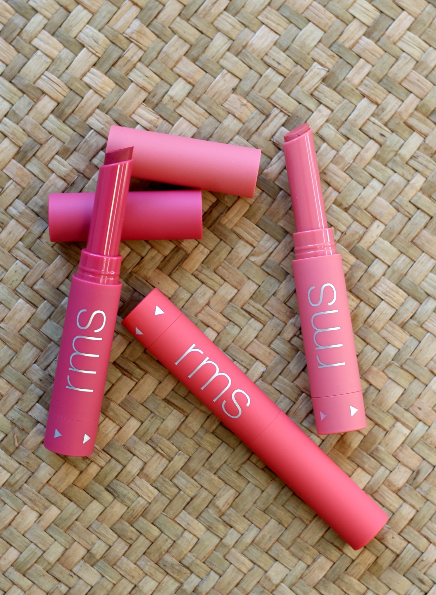 Four pink rms lipsticks arranged on a woven straw surface, with some tubes open and others closed.